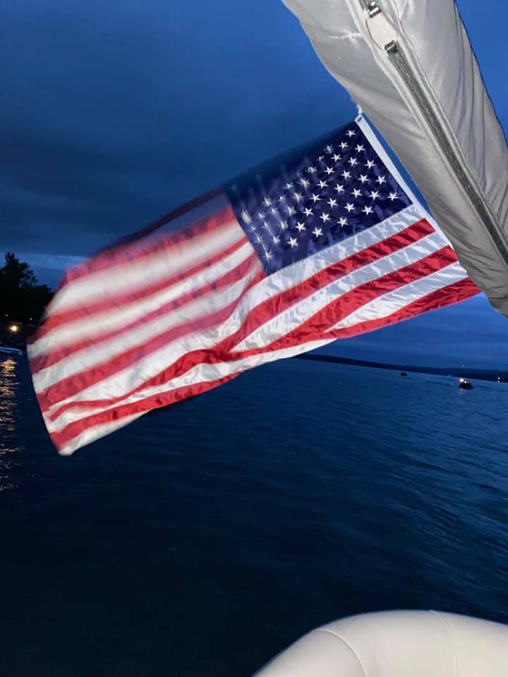 An American flag waves on a boat at dusk. The sky is dark with thick clouds, and calm water is visible in the background, with a few faint lights in the distance.