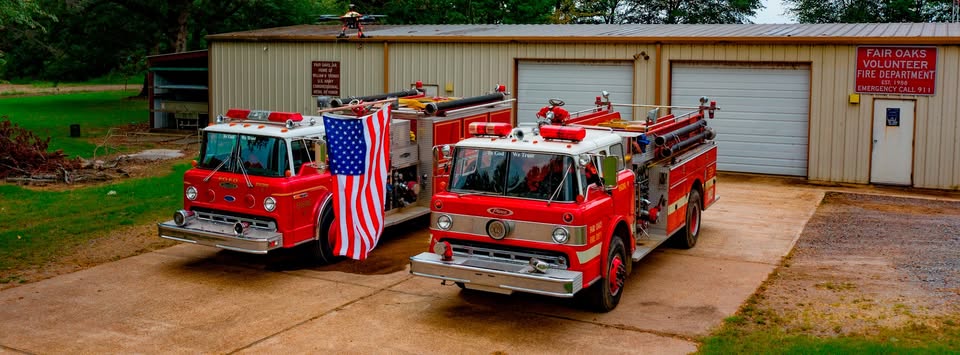 Two red fire trucks are parked on a concrete driveway in front of a beige metal building labeled Fair Oaks Volunteer Fire Department. An American flag is draped on the left truck. Trees are in the background.