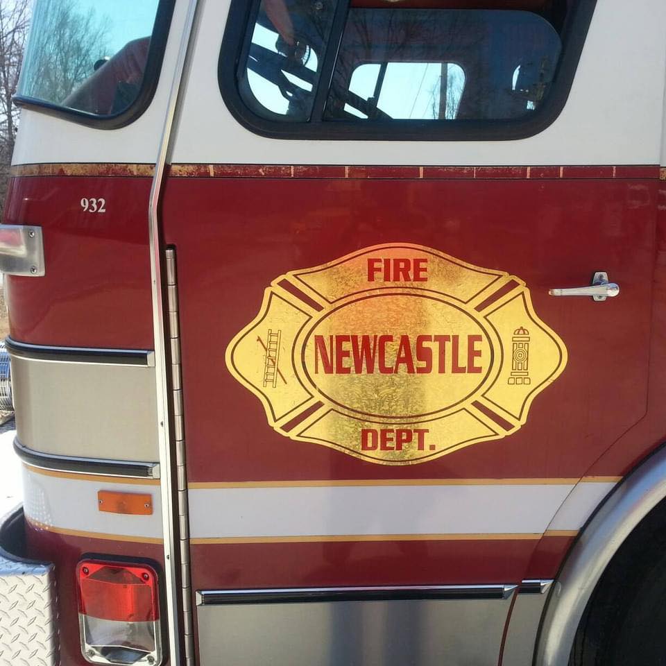 Side view of a red and white fire engine with Newcastle Fire Dept. in gold letters on the side, along with images of firefighting tools. The vehicle number 932 is visible above.