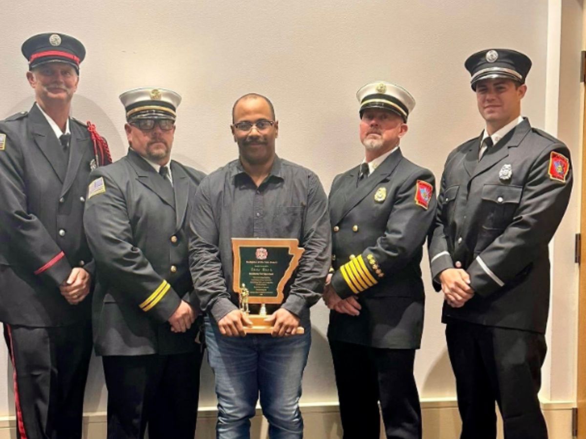 Five men stand together in front of a white wall. Four wear formal firefighter uniforms and hats, and one in the center, wearing glasses and a dark shirt, holds a wooden plaque with an award.
