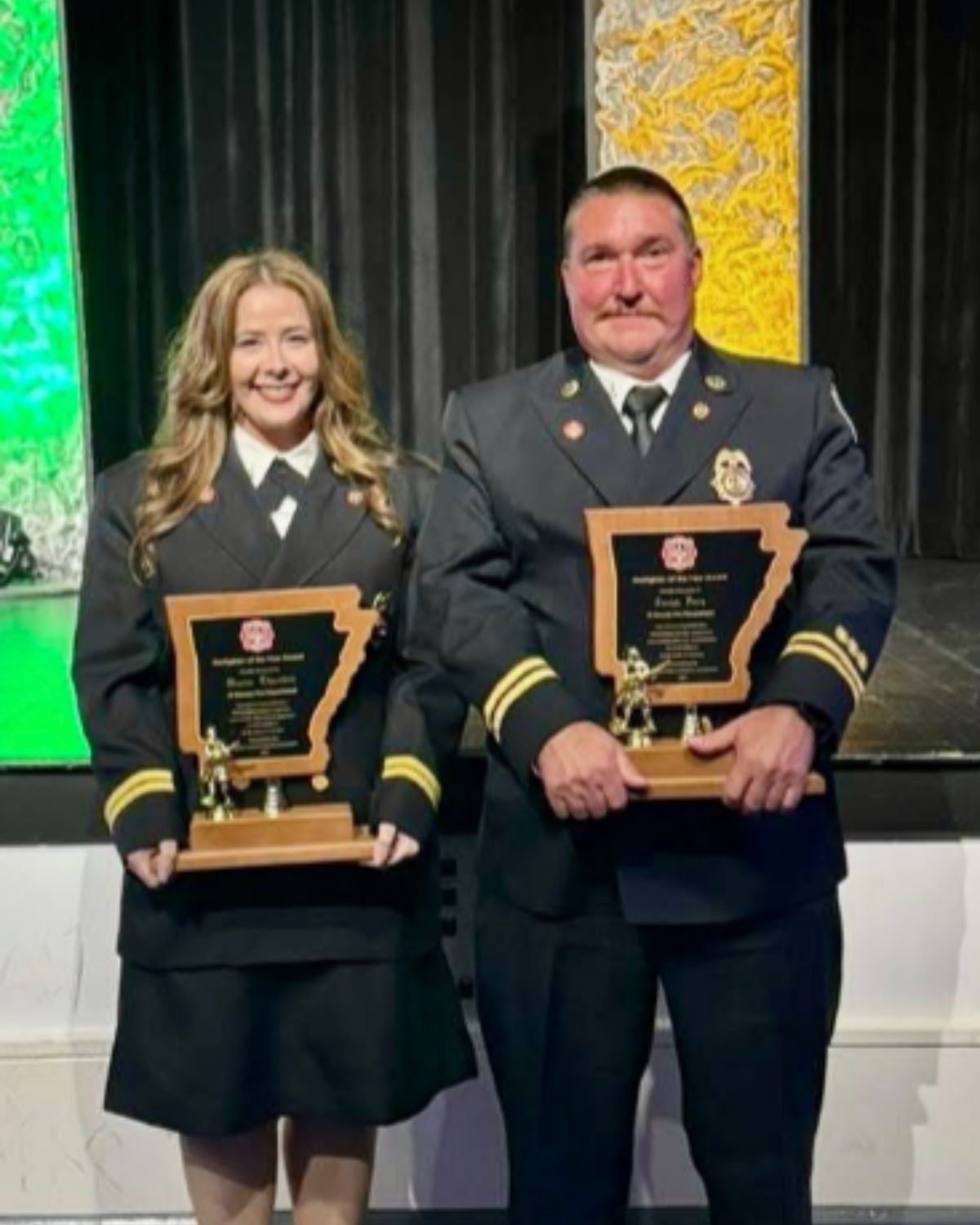 Two uniformed firefighters, one woman and one man, stand side by side on a stage holding matching plaques shaped like the state of Arkansas. They are smiling and dressed in formal attire.