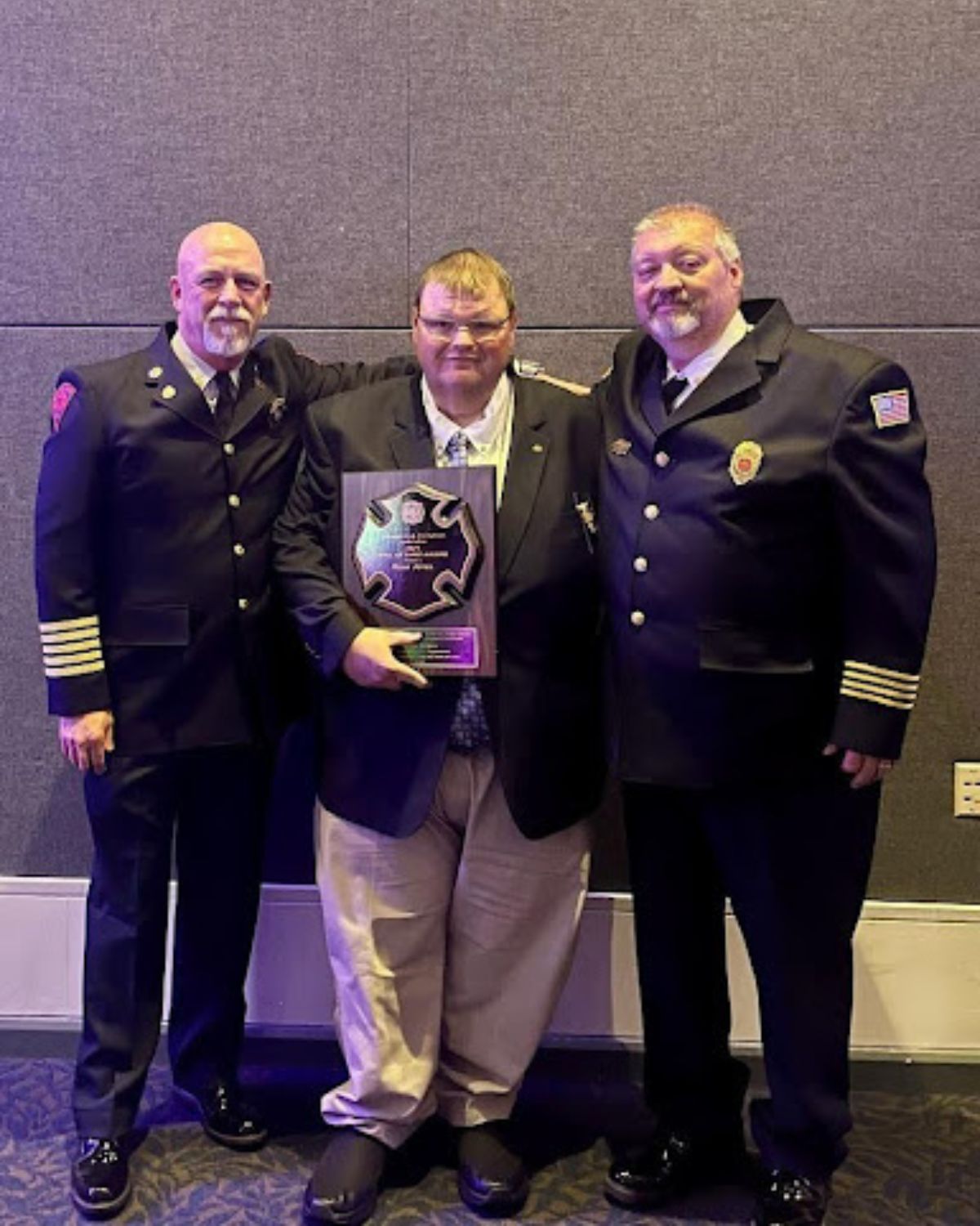 Three men in formal fire department uniforms pose together; the man in the center, wearing a suit, holds a plaque shaped like a fire department emblem. They stand against a gray wall at an indoor event.