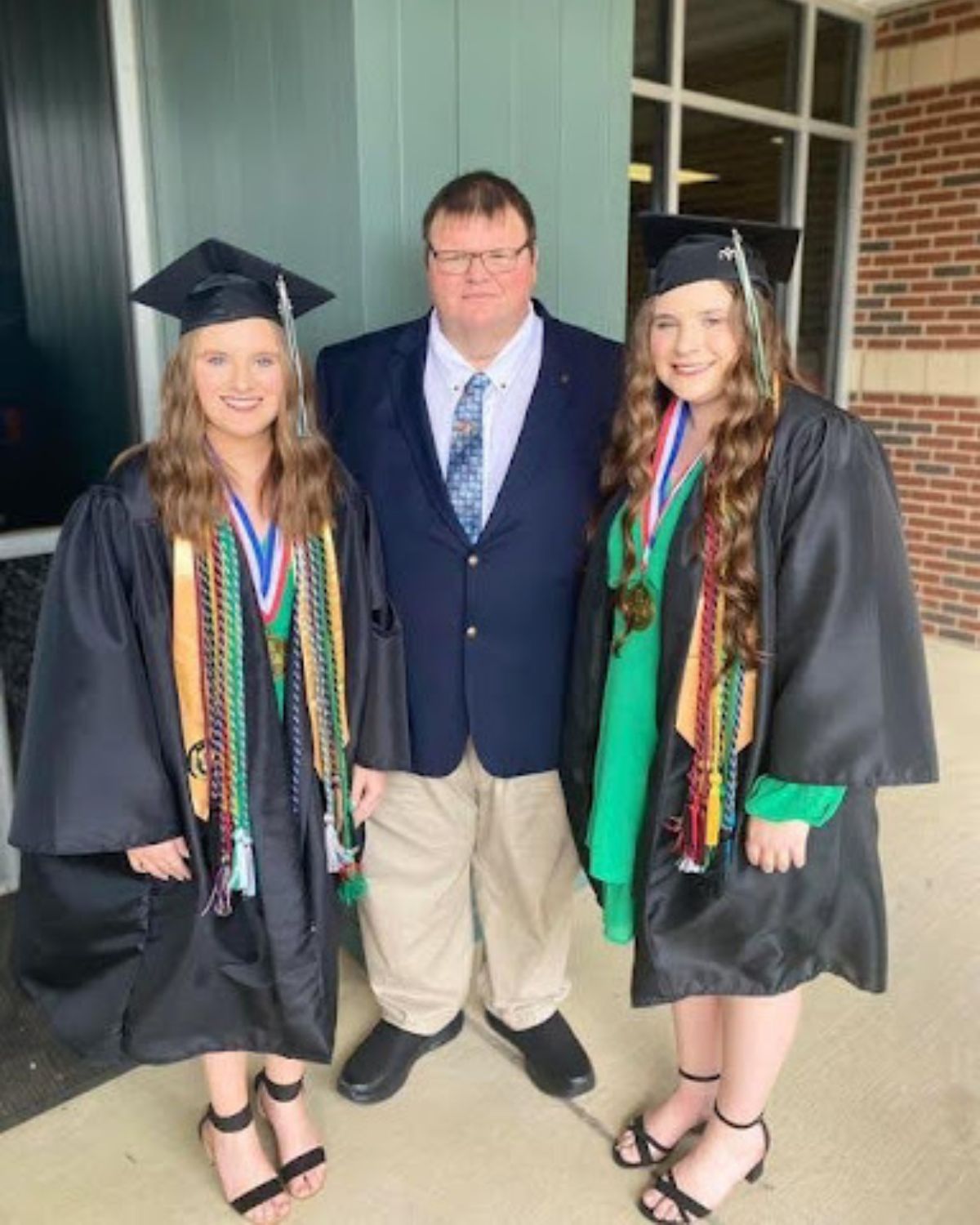 Two young women in graduation caps and gowns stand on either side of an older man in a suit and tie. All three are smiling and posing for a photo outside a building.
