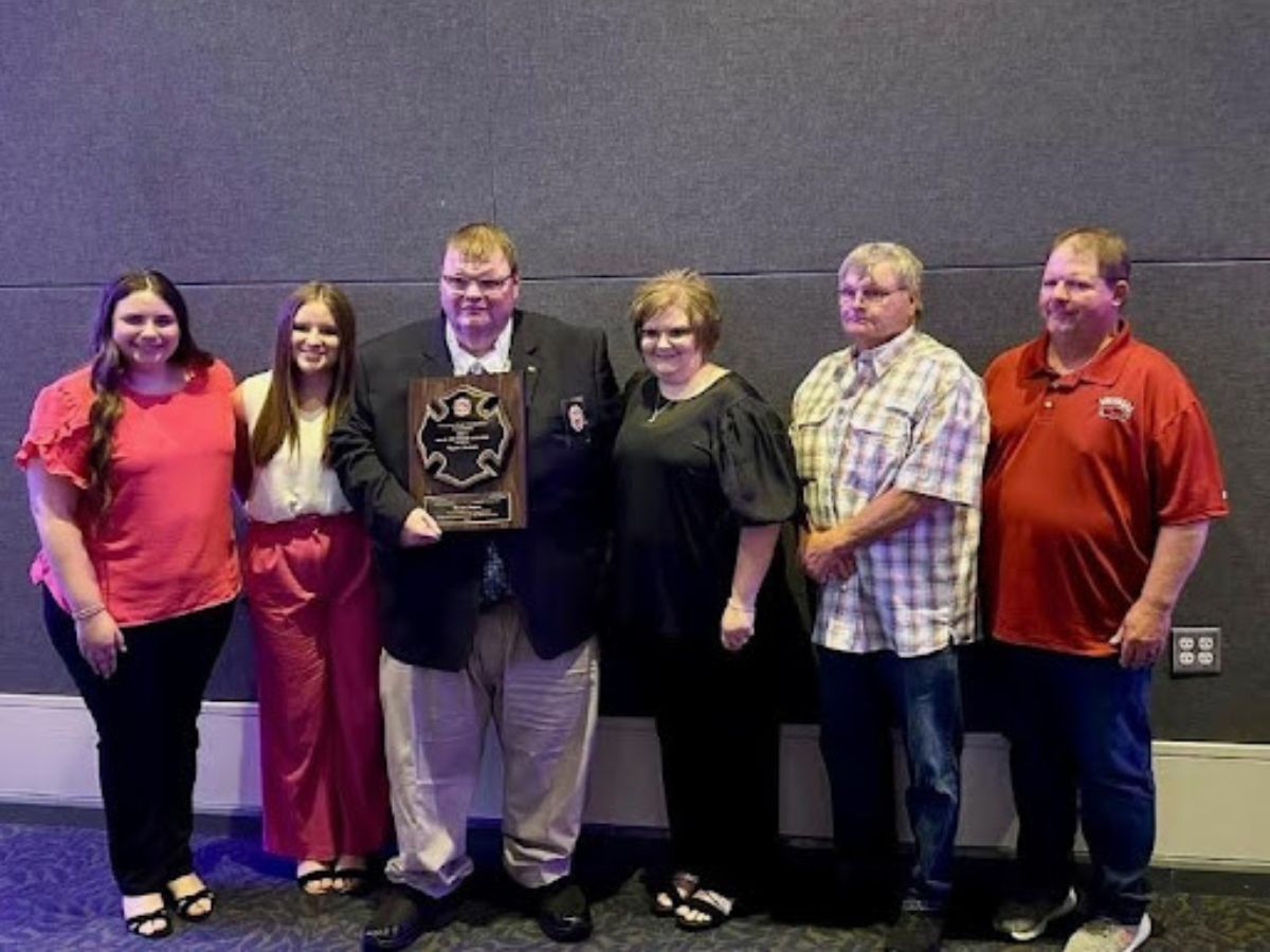 Six people stand side by side indoors; the man in the center holds a large plaque. They are all smiling and dressed in semi-formal attire, standing against a plain, dark wall.