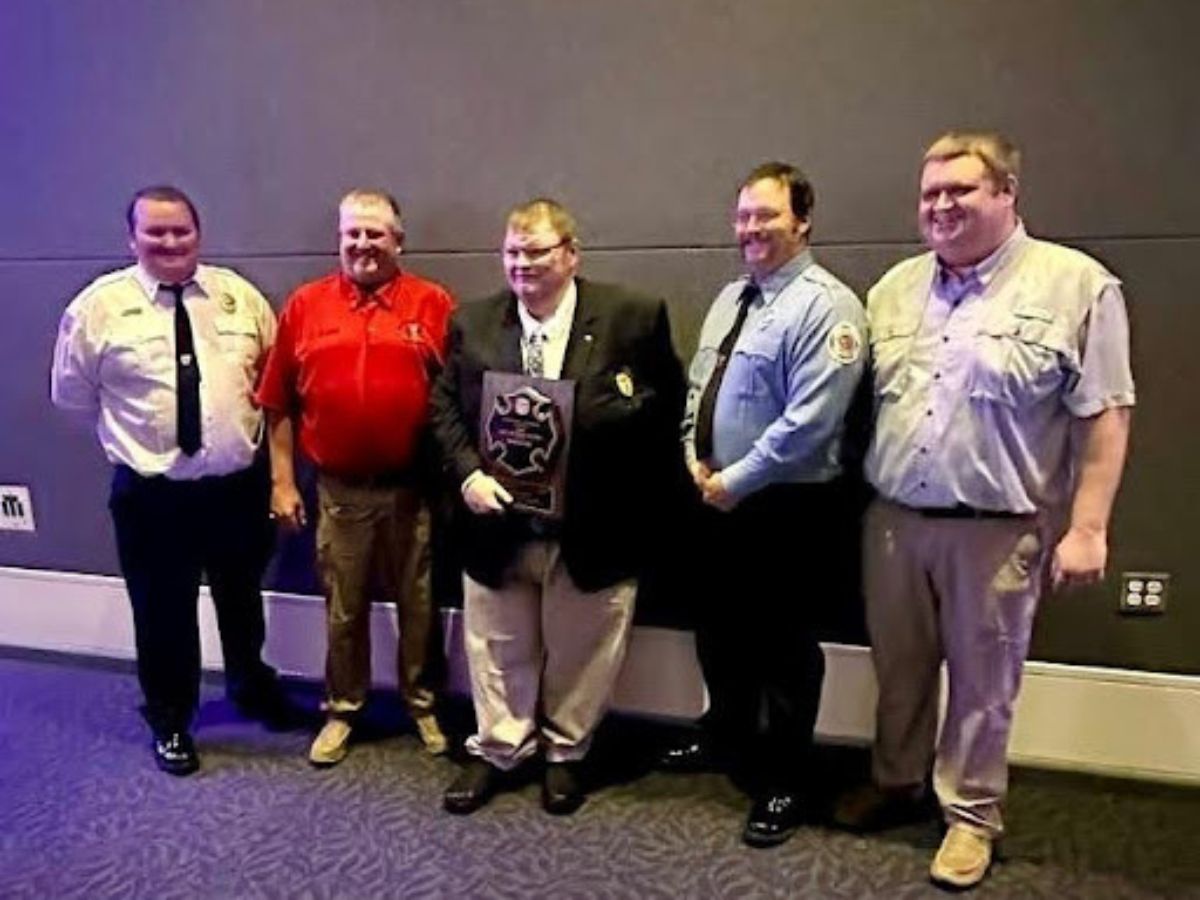 Five men stand indoors against a gray wall; one in the center holds a plaque. They are dressed in various uniforms and business attire, smiling and posing for a group photo on carpeted flooring.