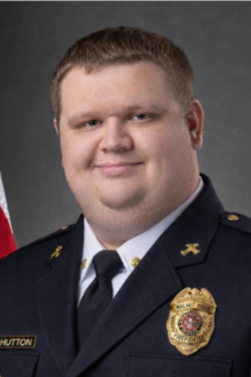 A man in a dark formal uniform with a badge and pins smiles at the camera. An American flag is partially visible in the background.