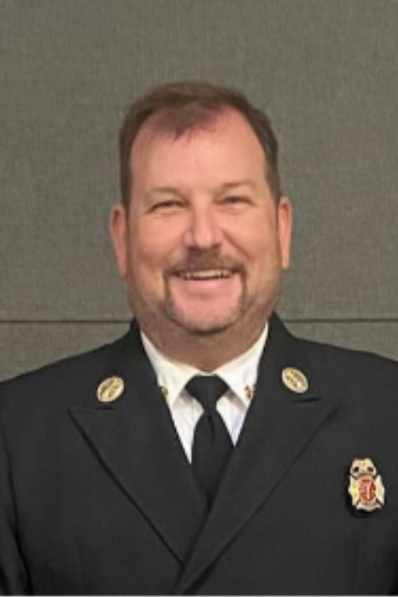 A man in a dark dress uniform with fire department badges on his collar and chest, standing in front of a plain gray background, smiling at the camera.