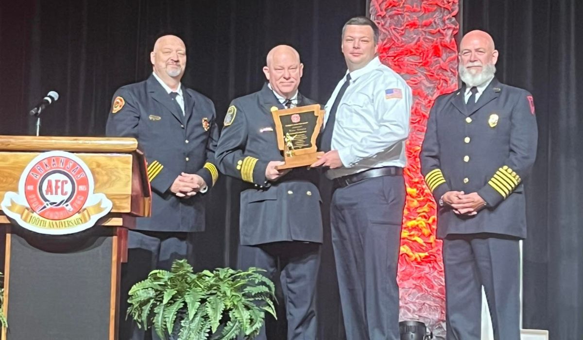2026 ASFFA Firefighter of the Year Blake Cupples 2 Four men in formal fire department uniforms stand on a stage. One man in a white shirt receives a plaque from another. A podium with the AFC logo and a potted plant are visible in the foreground.