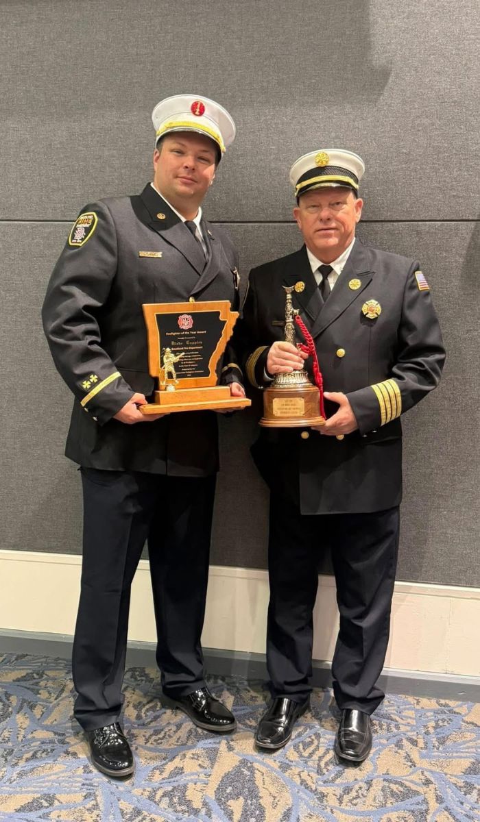 2026 ASFFA Firefighter of the Year Blake Cupples Two uniformed firefighters stand side by side indoors, each holding a large award. One has a wooden plaque with a gold firefighter figure, and the other holds a trophy topped with a fire helmet. Both are smiling.