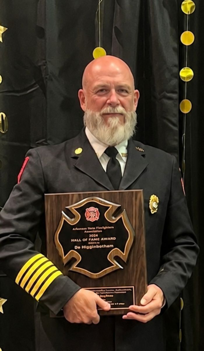 Bo Higginbotham - Hall of Fame 2026 A man in a formal fire chief uniform holds a large plaque reading Arkansas State Firefighters 2024 Hall of Fame Award in front of a black and gold decorated backdrop.