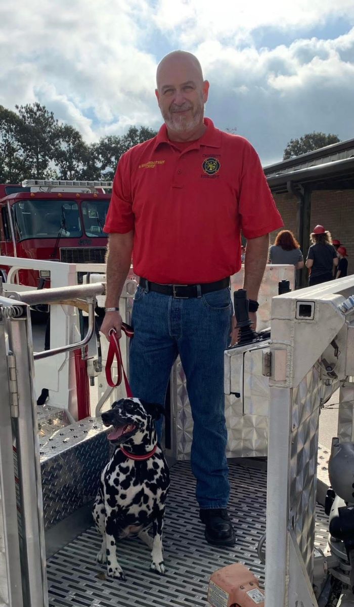 Bo and Ember his Dalmatian A smiling man in a red fire department shirt and jeans stands on a fire truck platform, holding a red leash attached to a sitting Dalmatian dog. A fire truck and several people are visible in the background.