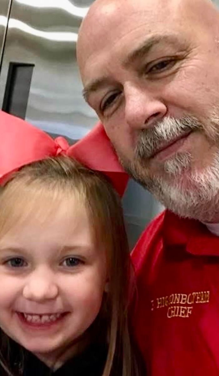 Bo and his granddaughter A smiling young girl with a large red bow poses for a selfie with a bearded man wearing a red shirt that says CHIEF. They are indoors, and both are looking at the camera.
