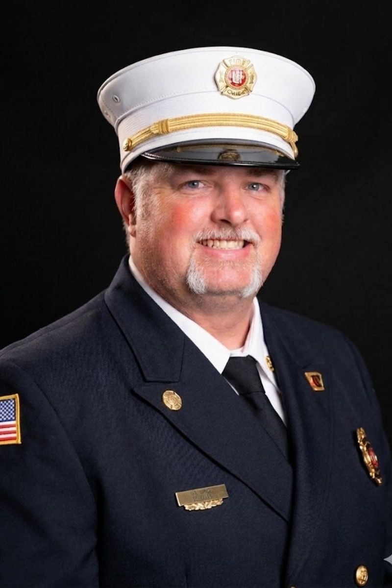 Brad Moore - New A smiling man in a fire chief’s dress uniform with a white hat, gold badge, and various pins, including an American flag patch on the shoulder, poses against a black background.