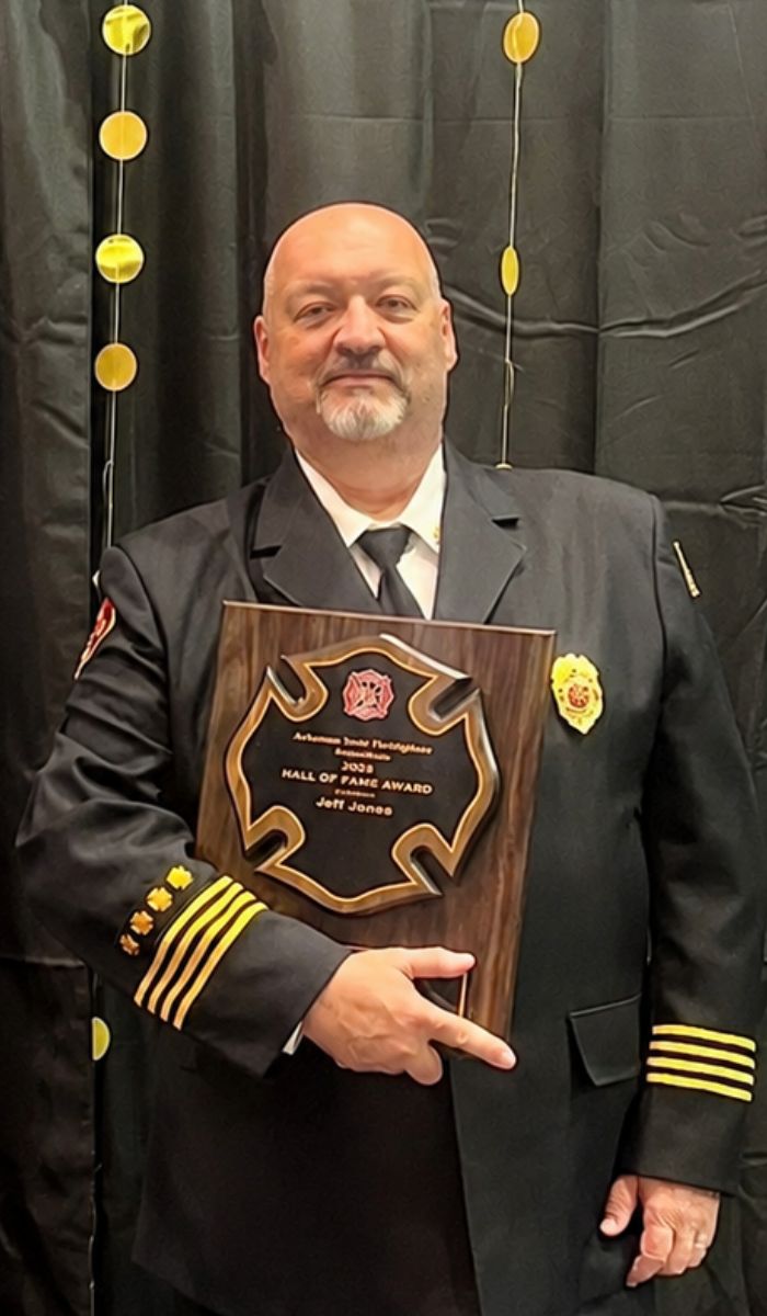 Jeff Jones - Hall of Fame 2026 A man in a formal firefighter’s uniform holds a large plaque that reads “2023 Hall of Fame Award, Jeff Jones” with a fire department emblem, standing in front of a black curtain decorated with gold circles.