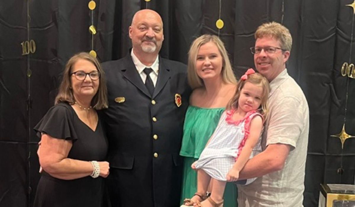 Jeff and his family Five people pose together in front of a black backdrop with gold decorations. One man is wearing a dark suit with a badge, while the others are dressed in casual and semi-formal attire. A child in a dress is held by a man on the right.