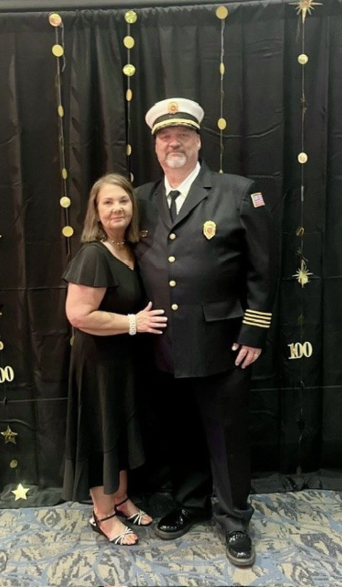 Jeff and his wife A woman in a black dress stands beside a man in a fire chief uniform with a hat, in front of a black backdrop decorated with gold stars and 100 signs. They are smiling and posing together.