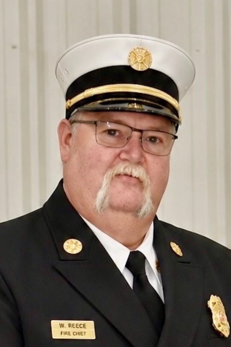 Wayne Reece - Brookland Fire Protection District A man with a white mustache wears a fire chief’s uniform, including a black coat with gold buttons, a white hat with a gold badge, and glasses. He stands in front of a light-colored, striped background.