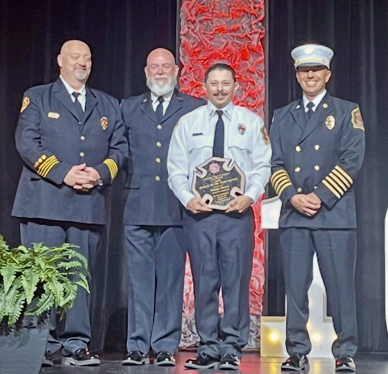 Screenshot Four men in formal fire department uniforms stand on stage. The man second from the right holds a plaque and smiles. A potted fern sits on the left, and a red-lit textured backdrop is behind them.